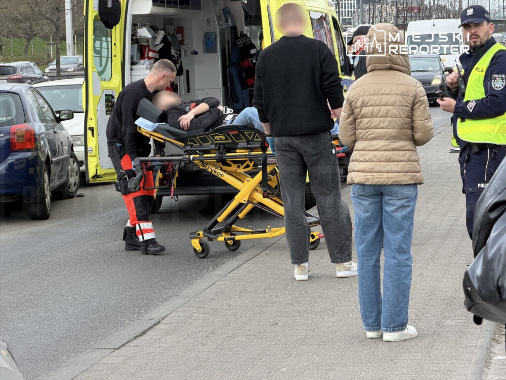 Mężczyzna w mundurze medycznym pomaga osobie leżącej na noszach obok karetki, a w tle obserwują ich przechodnie.