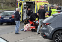 Ratownicy medyczni udzielają pomocy osobie leżącej na chodniku obok karetki, w tle widoczne samochody.