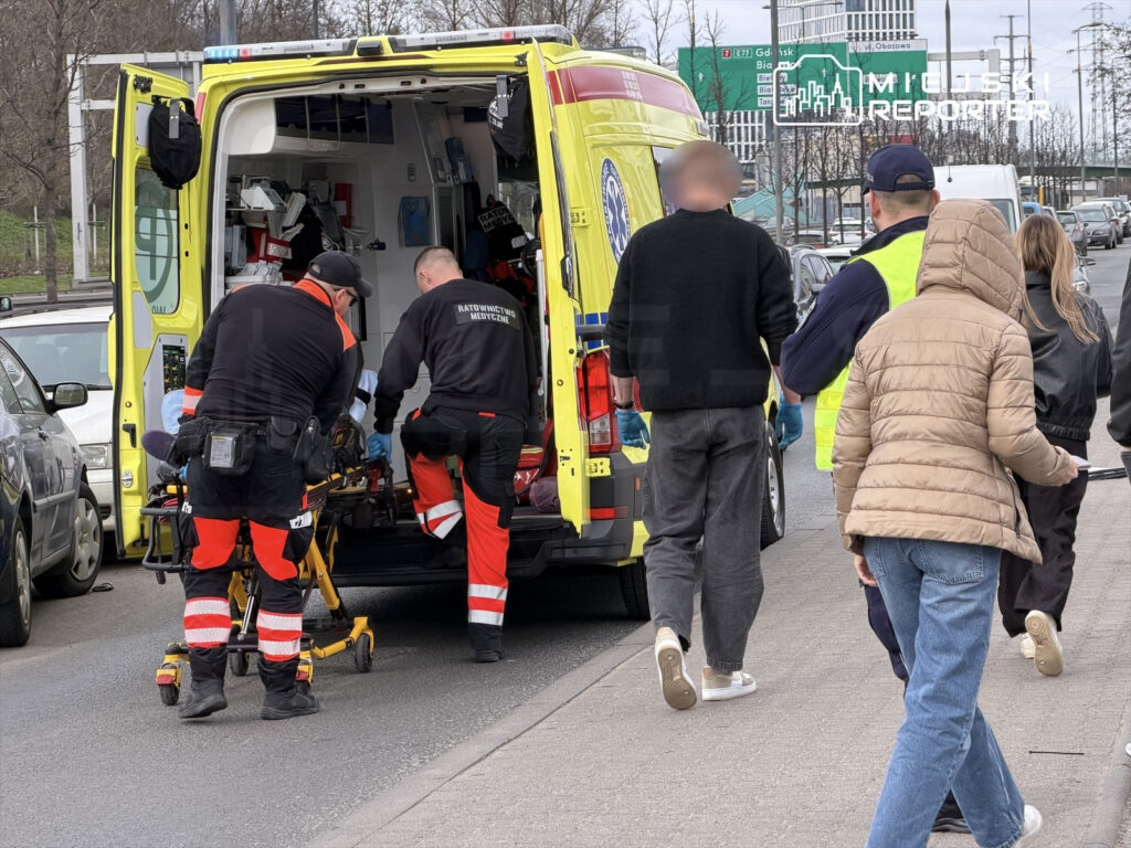 Ratownicy medyczni przenoszą pacjenta na noszach do karetki, podczas gdy przechodnie obserwują sytuację na ulicy.