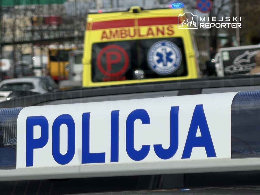 Policyjny radiowóz z widocznym napisem "POLICJA" na dachu, w tle karetka pogotowia oraz ruch uliczny.