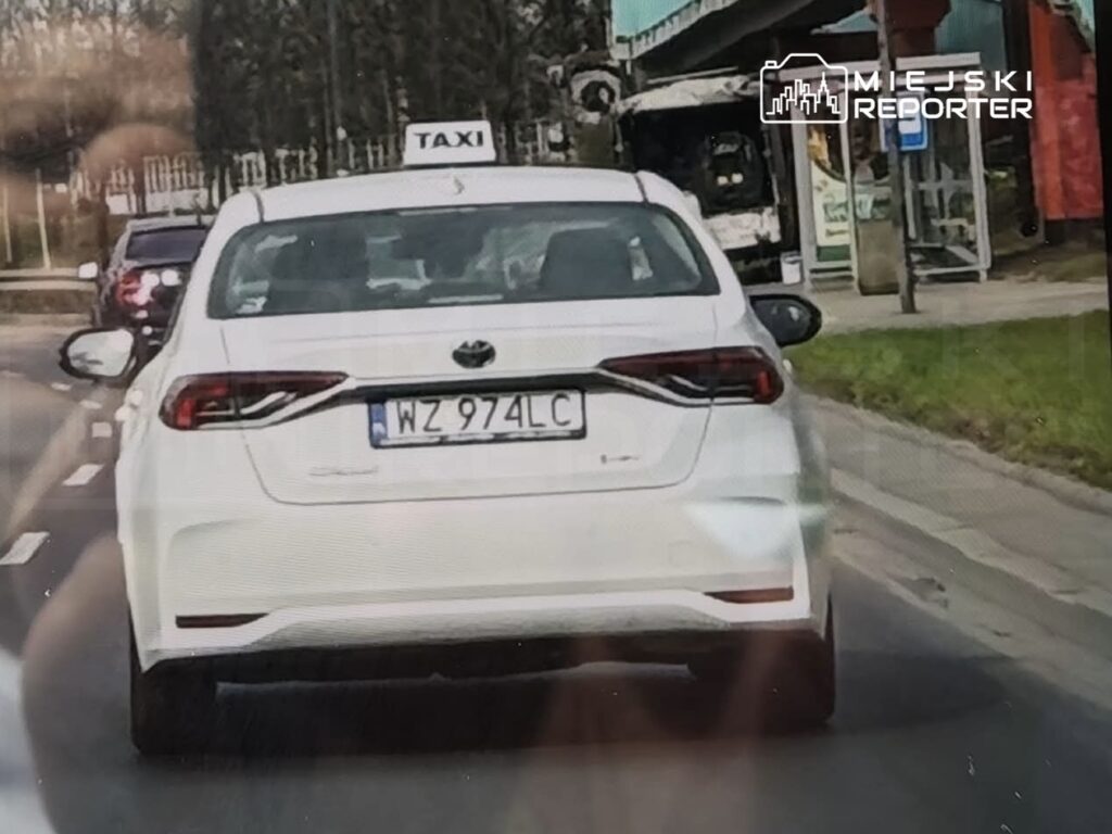 Biały samochód osobowy z oznaczeniem "TAXI" na dachu porusza się po ulicy.