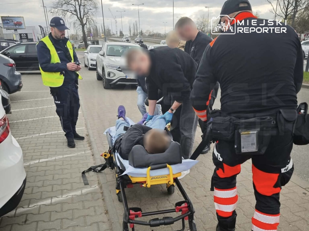 Grupa ratowników medycznych udziela pomocy mężczyźnie leżącemu na noszach w strefie parkingowej.