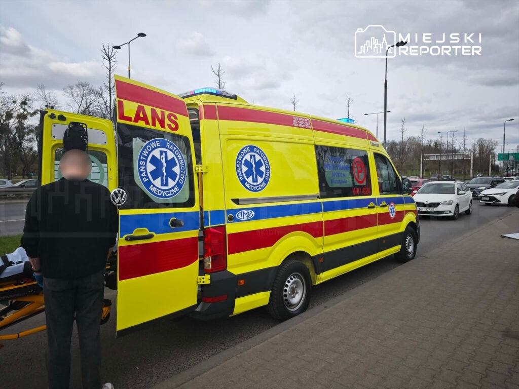 Mężczyzna w czarnej bluzie stoi obok otwartej karetki pogotowia, na drodze w miejskim otoczeniu.