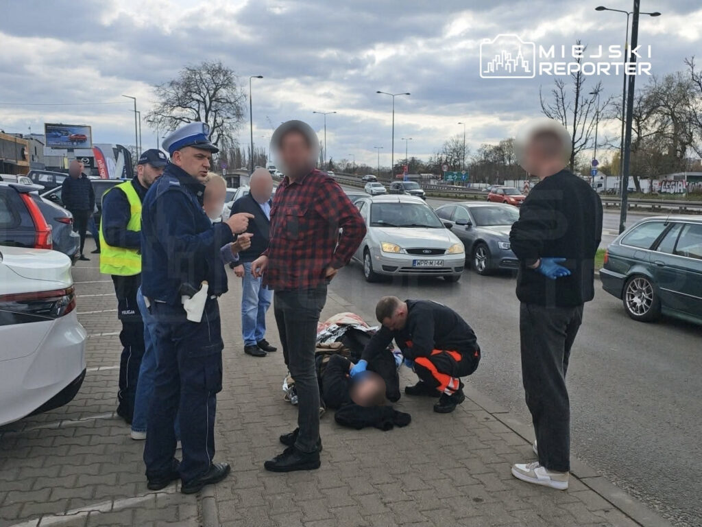 Policjant w mundurze rozmawia z mężczyzną w kratkę, podczas gdy ratownik medyczny udziela pomocy osobie leżącej na chodniku.