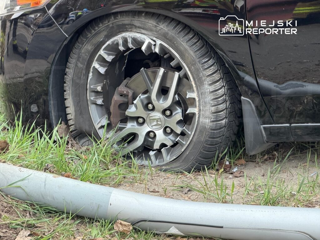 Samochód z uszkodzoną oponą leży na poboczu drogi, obok trawy i rury.