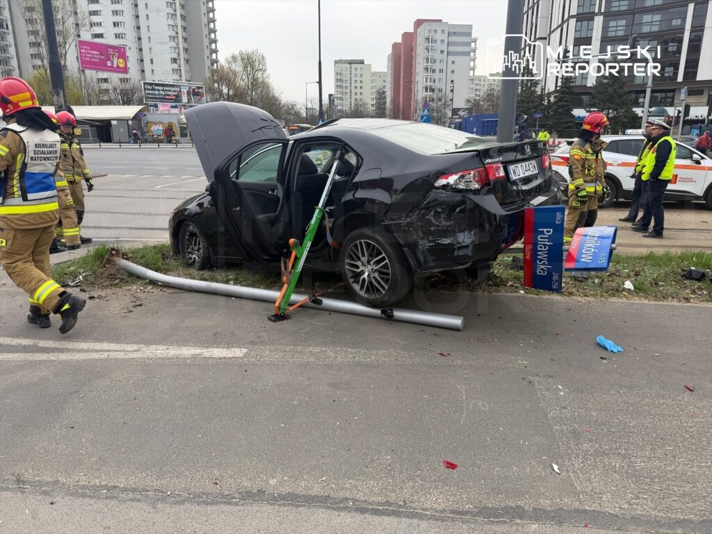 Czarny samochód osobowy uderzył w słup, a obok pracują strażacy w mundurach ratunkowych. Na drodze leżą zniszczone elementy.