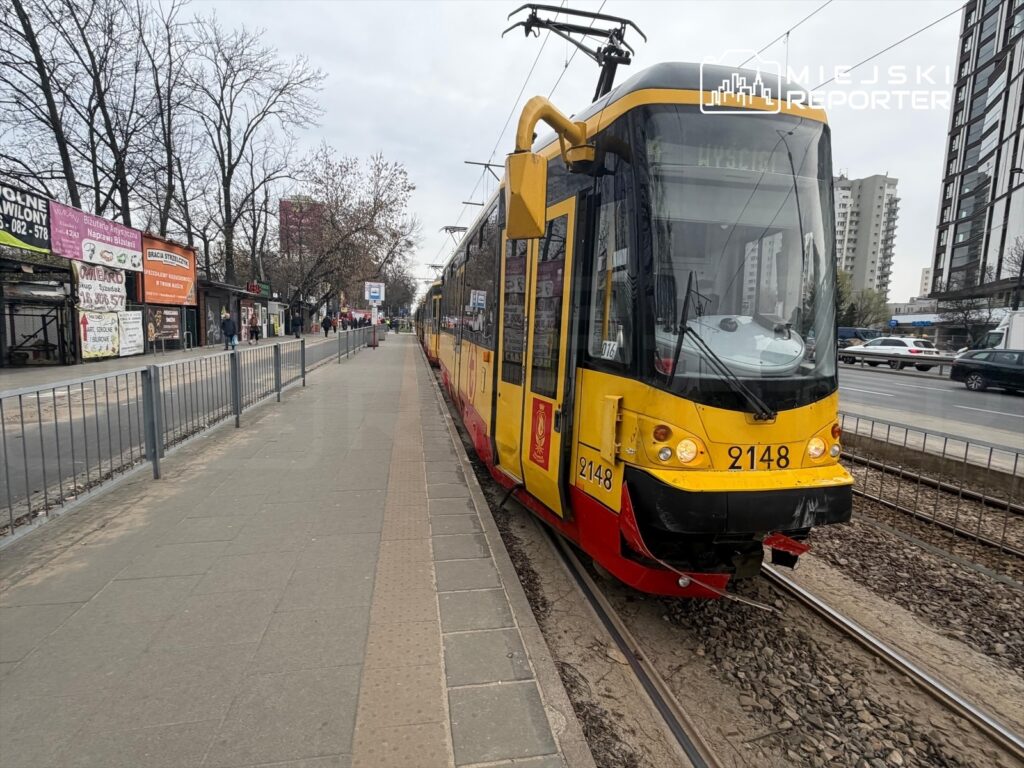 Tramwaj o numerze 2148 zatrzymany na przystanku, obok pasażerowie czekają na przyjazd. W tle widoczne sklepy i budynki.