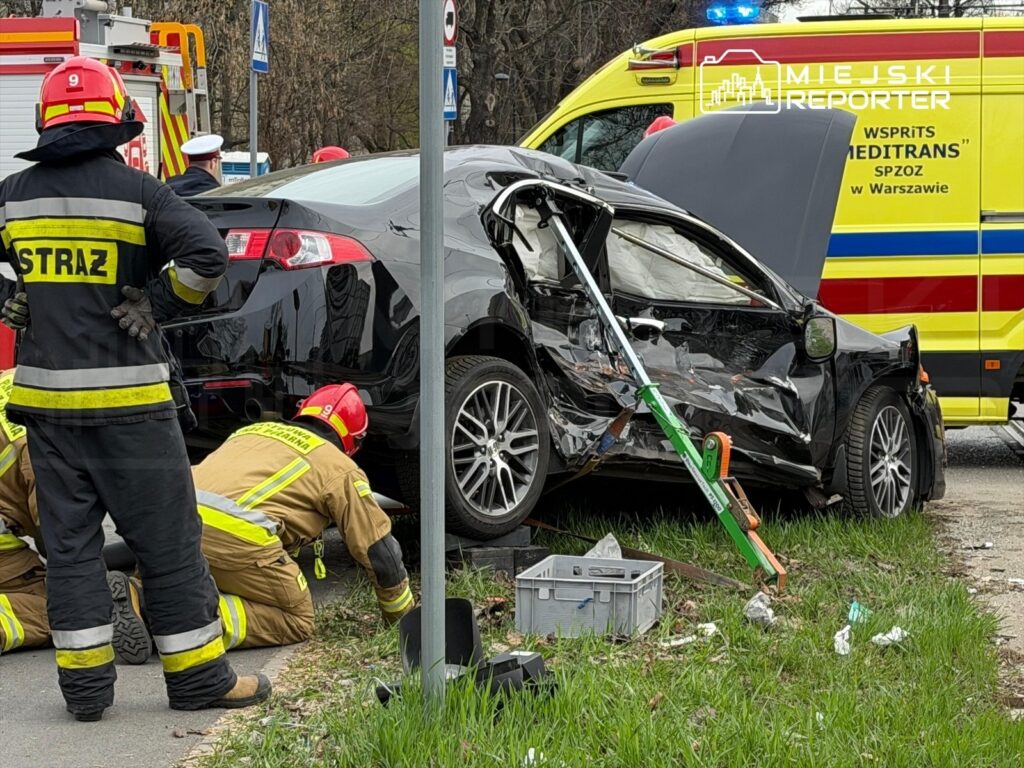 Strażacy w mundurach pracują przy uszkodzonym czarnym samochodzie osobowym na poboczu drogi, obok karetki pogotowia.