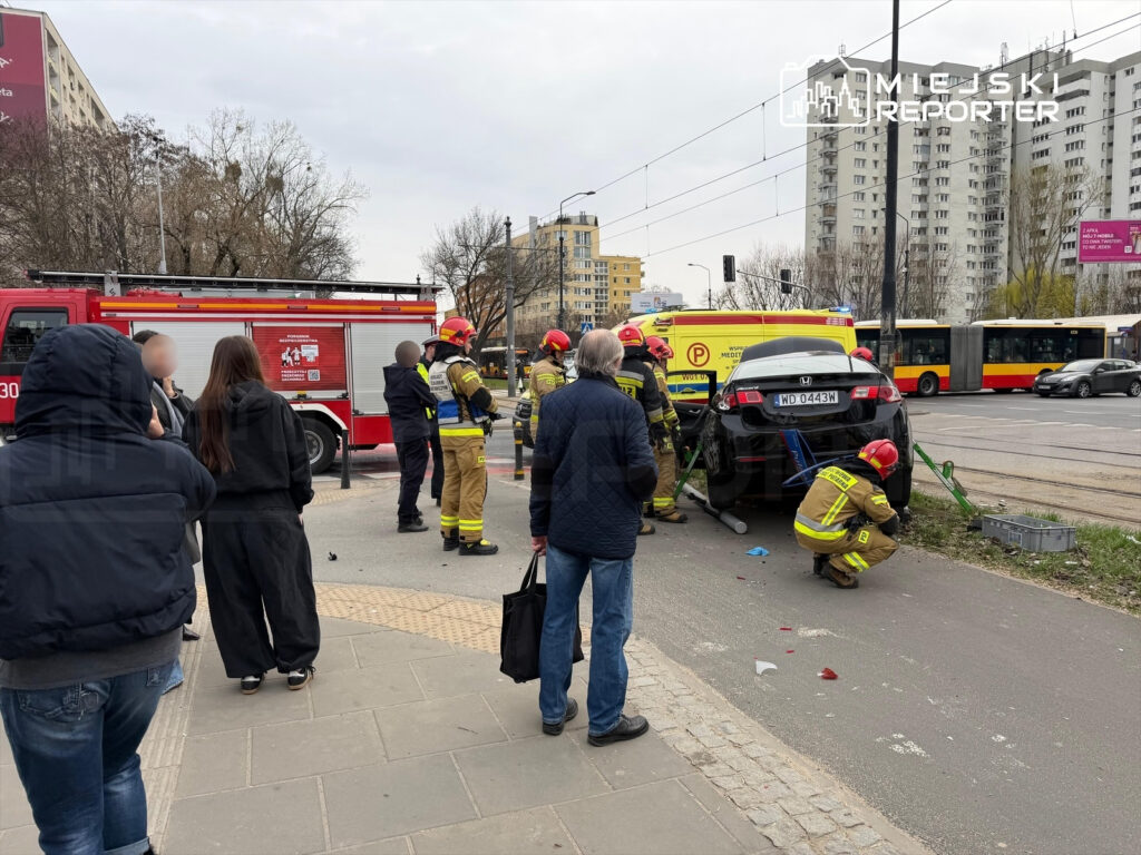 Grupa strażaków zabezpiecza miejsce wypadku, gdzie czarny samochód jest podnoszony z jezdni, a obok stoją gapie.