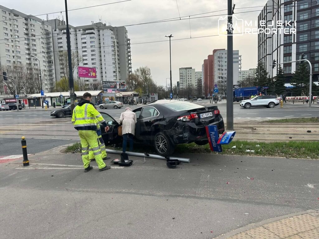 Kobieta sprawdzająca uszkodzenia czarnego samochodu osobowego, obok stoi mężczyzna w odblaskowym uniformie.