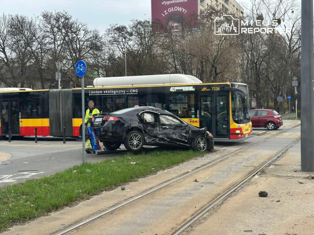 Czarny samochód osobowy z uszkodzoną boczną częścią stoi na torach tramwajowych obok przystanku, obok autobusu komunikacji miejskiej.