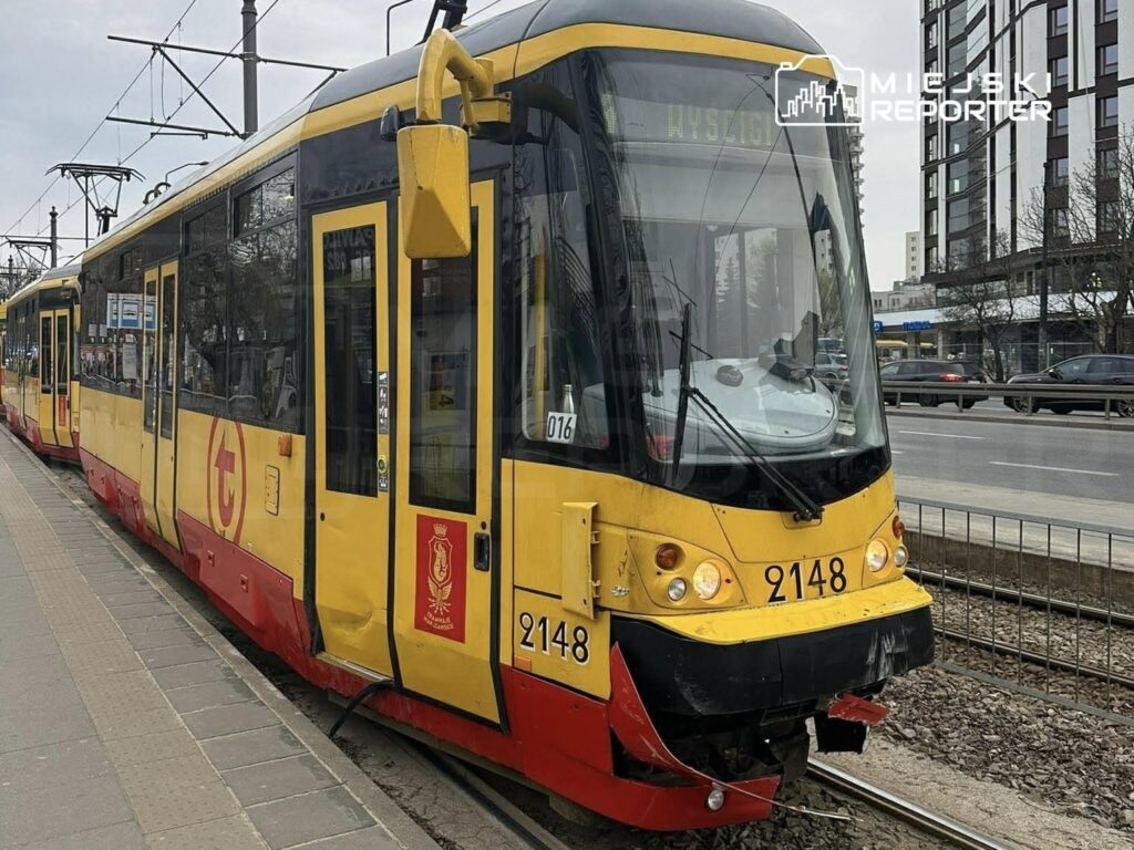 Tramwaj w kolorze żółto-czerwonym zatrzymany na przystanku, obok ruchliwa ulica i nowoczesne budynki.