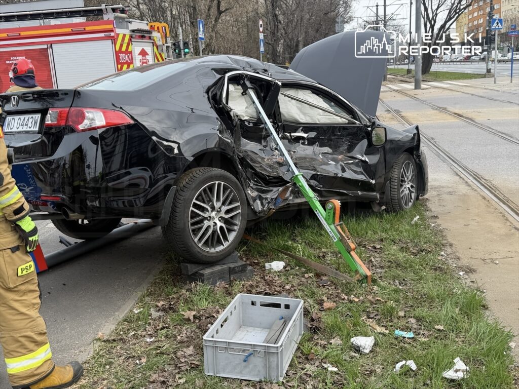 Czarny samochód osobowy z uszkodzoną karoserią stoi na trawie obok ulicy, w tle widać wóz strażacki i tramwajowe tory.