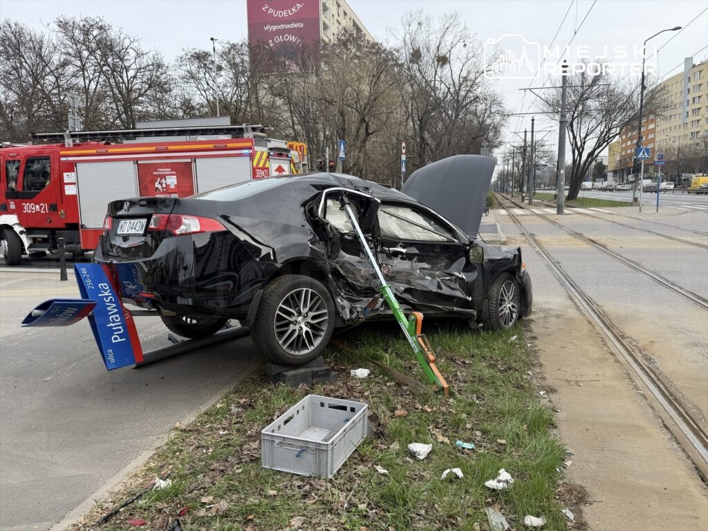 Zniszczony czarny samochód osobowy stoi na trawniku obok torów tramwajowych, a w tle widać wóz strażacki.