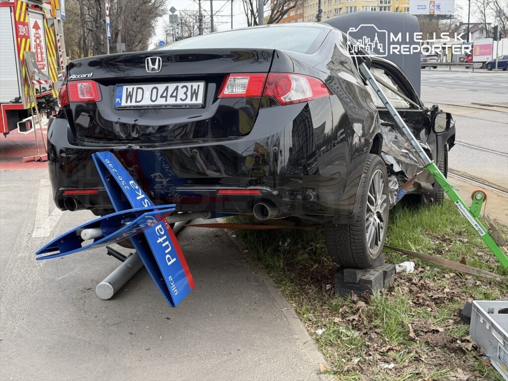Czarny samochód osobowy z uszkodzoną tylną częścią stoi na chodniku, obok widać sprzęt ratunkowy.