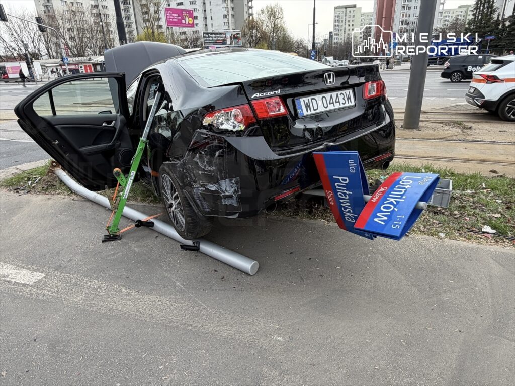 Czarny samochód osobowy z uszkodzeniami, stojący na poboczu drogi, z otwartymi drzwiami i z zamocowanym znakiem drogowym.