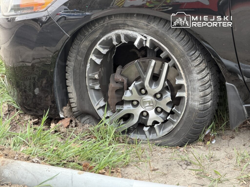Czarny samochód z uszkodzoną oponą stoi na poboczu, otoczony trawą i liśćmi.