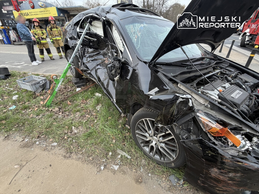 Czarny samochód osobowy z uszkodzoną boczną częścią stoi na trawie obok drogi, a obok niego dwóch strażaków w mundurach.