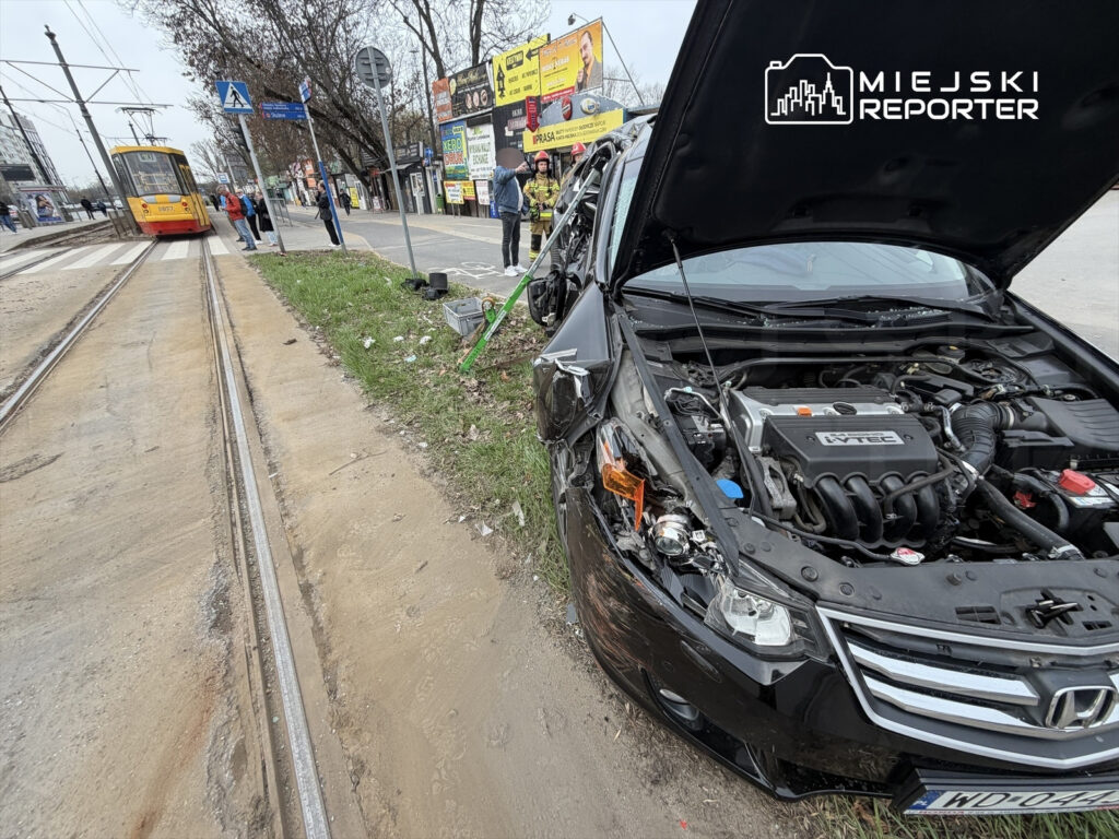 Samochód osobowy z uszkodzoną przednią częścią stoi na poboczu obok torów tramwajowych, w tle tramwaj i przechodnie.