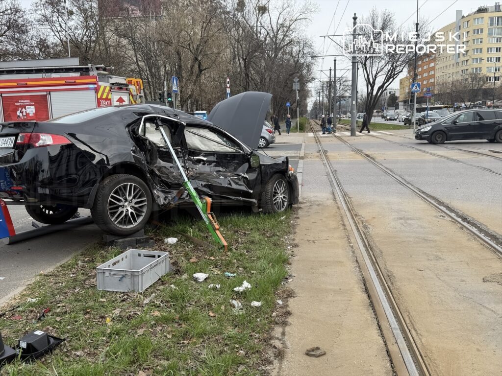 Zniszczony czarny samochód osobowy stoi na poboczu drogi, obok widać straż pożarną i przechodniów.