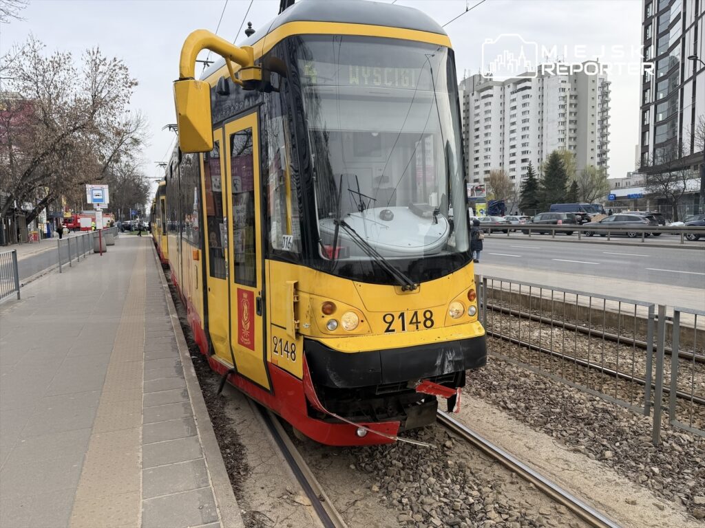 Tramwaj linii 2148 zatrzymany na przystanku, obok ruchliwej ulicy z samochodami i budynkami w tle.