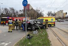 Grupa strażaków pracuje przy zderzonym samochodzie osobowym na ruchliwej ulicy, obok wozów strażackich i karetki.