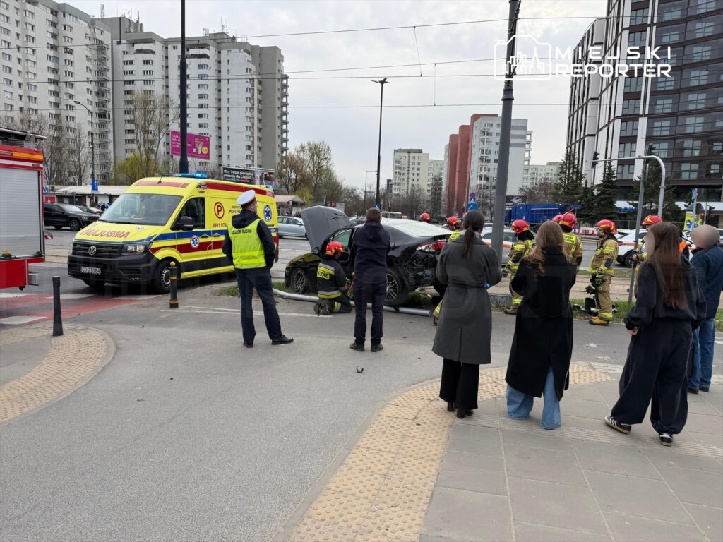 Grupa strażaków oraz ratowników medycznych udziela pomocy po wypadku samochodowym w miejskim otoczeniu, obok bloku mieszkalnego.