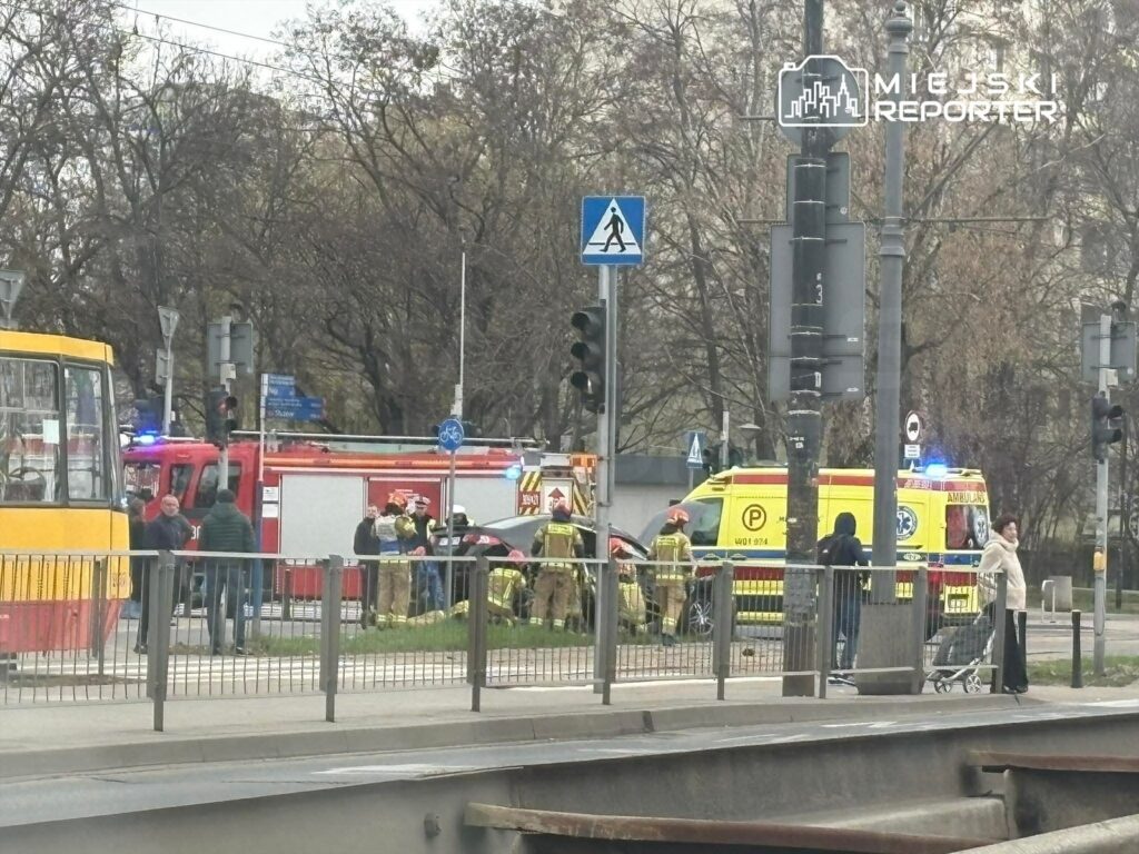 Grupa strażaków udziela pomocy poszkodowanemu na miejscu zdarzenia, obok wozów strażackich i karetki.