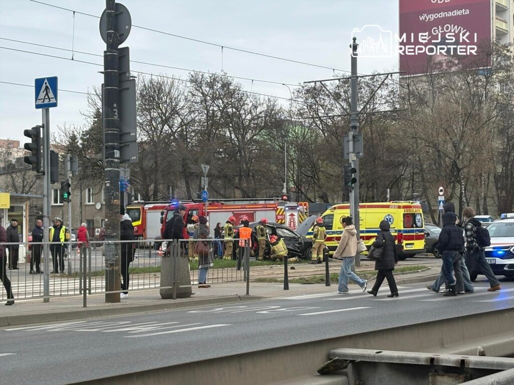 Na skrzyżowaniu ulic, grupa ludzi obserwuje akcję ratunkową po wypadku drogowym, w tle widoczne pojazdy ratunkowe.