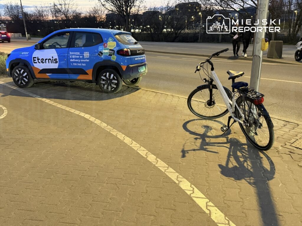 Na chodniku stoi biały rower zaparkowany obok niebieskiego samochodu dostawczego z reklamą firmy Eternis.