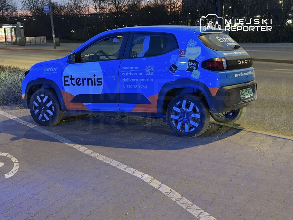 Niebiesko-pomarańczowy samochód dostawczy zaparkowany na chodniku, z widocznym napisem "Eternis" oraz informacjami o usługach dostawczych.