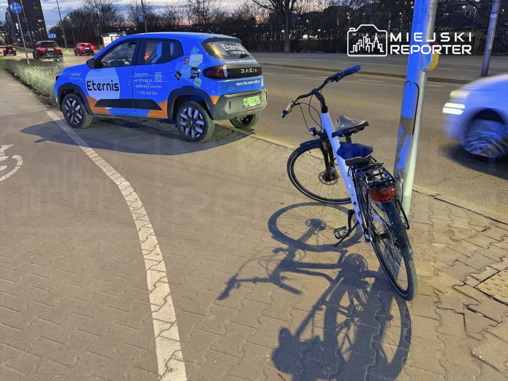 Rower zaparkowany przy krawężniku obok samochodu dostawczego z reklamą, w tle widoczne ruchliwe ulice.