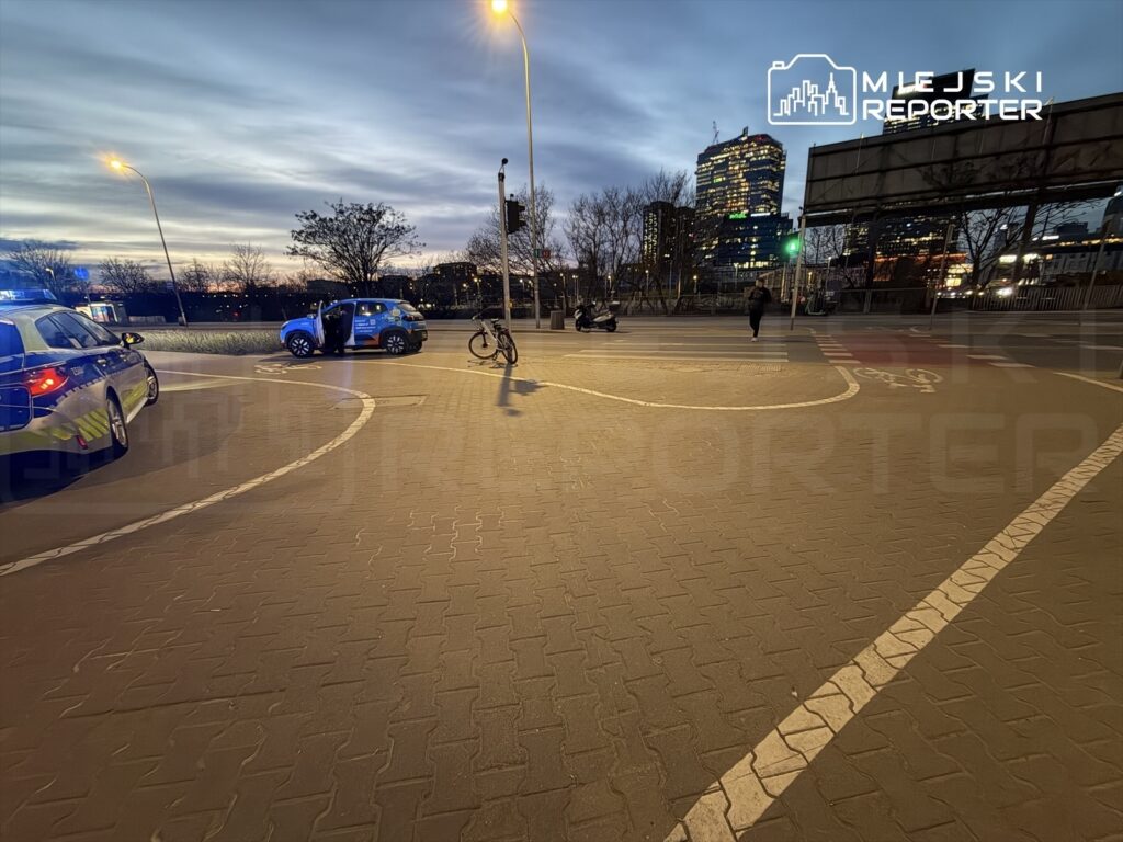 Policyjny radiowóz z włączonymi sygnałami świetlnymi stoi na skrzyżowaniu, obok rower i mężczyzna przechodzący przez ulicę.