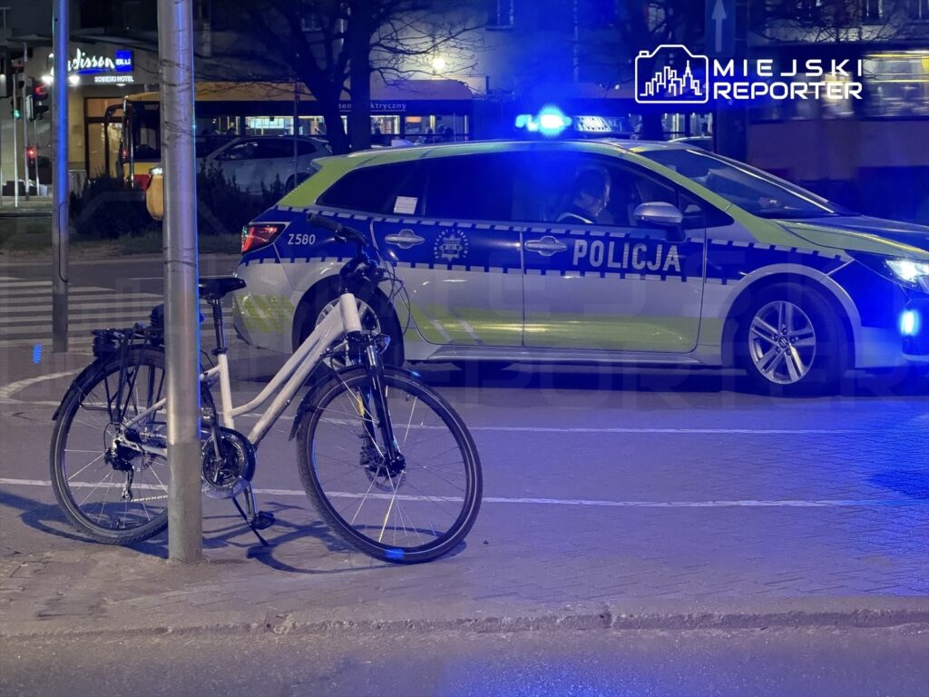 Na ulicy stoi biały rower przyparty do słupa, obok zaparkowany radiowóz policyjny z włączonymi sygnałami świetlnymi.