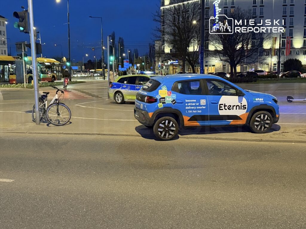 Na skrzyżowaniu stoi niebieski samochód dostawczy z logo "Eternis", obok zaparkowany rower. W tle widoczny ruch uliczny.