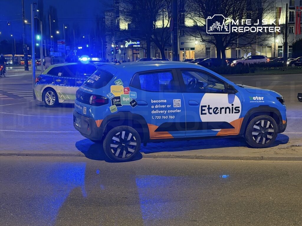 Niebieski samochód dostawczy z logo Eternis zatrzymany na ulicy, obok radiowozu policyjnego z włączonymi sygnałami świetlnymi.