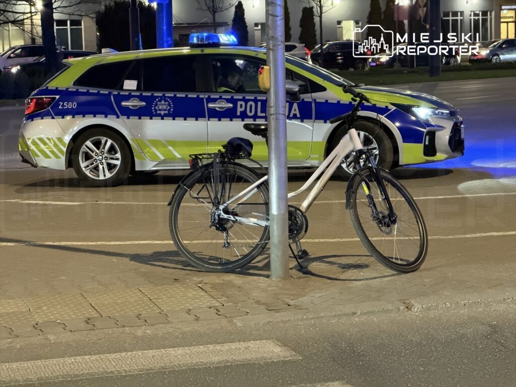 Rower zaparkowany obok słupa, a w tle widoczny radiowóz policyjny z włączonymi światłami.