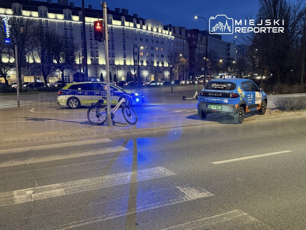 Rower stoi przy sygnalizatorze świetlnym, obok zaparkowanej policyjnej radiowozu oraz samochodu firmowego.