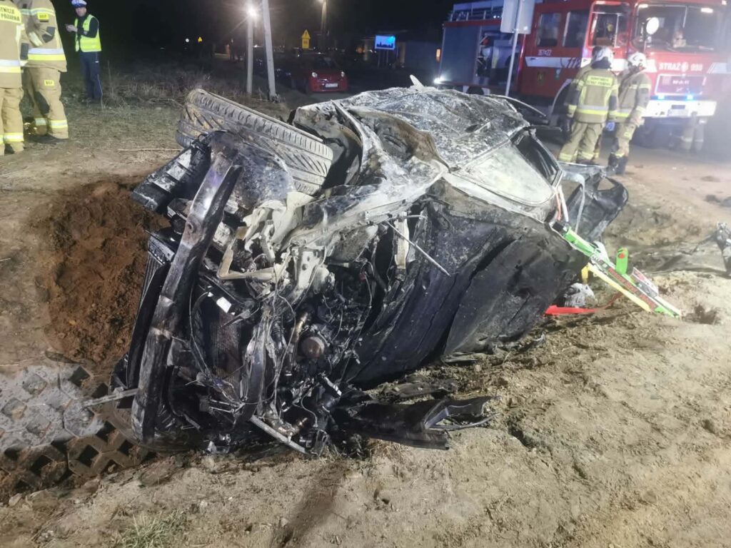 Uszkodzony samochód leży na boku w pobliżu drogi, a obok pracują strażacy w mundurach.