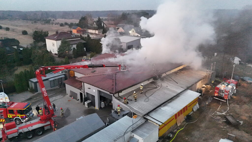 Strażacy gaszą pożar na dachu budynku przemysłowego, z którego unosi się dym, w otoczeniu zabudowy mieszkalnej.