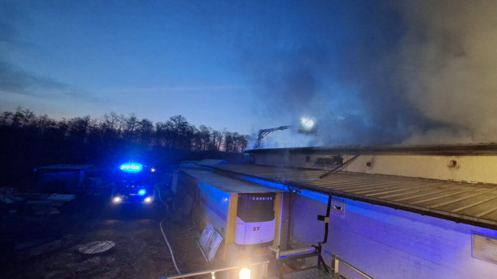 Strażacy gaszą pożar dachu budynku, używając podnośnika, w otoczeniu ciemnego nieba i zaparkowanych pojazdów.