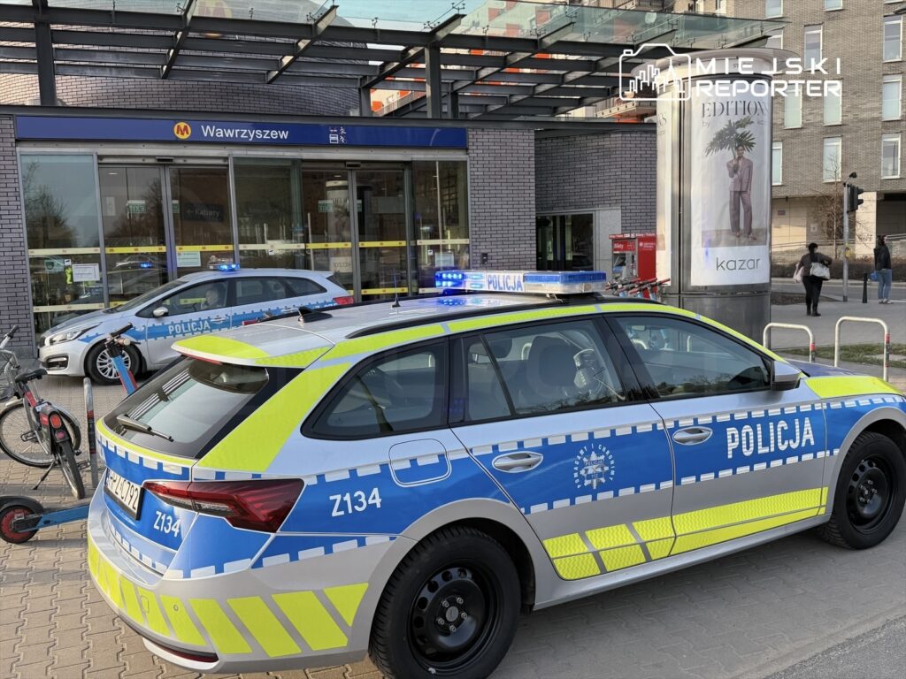 Policyjne samochody z włączonymi sygnałami świetlnymi stoją przed stacją metra, w tle widać przechodniów i rowery.