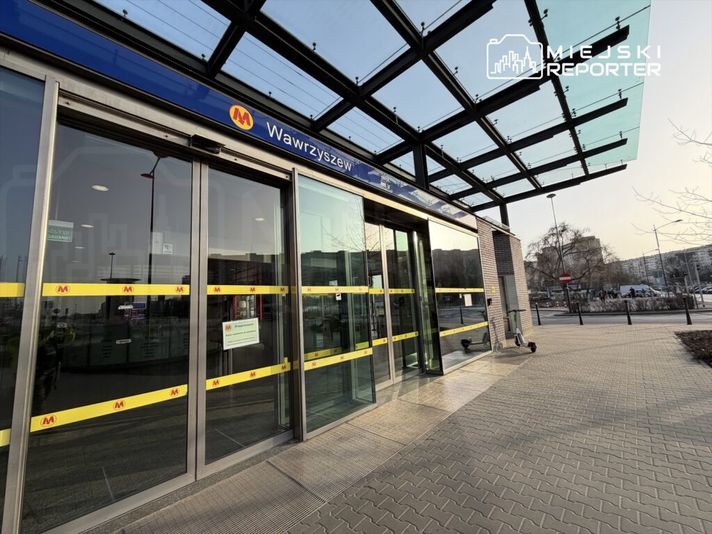 Nowoczesna stacja metra z przeszklonym dachem, szklane drzwi z żółtymi taśmami, widok na chodnik i otoczenie miejskie.