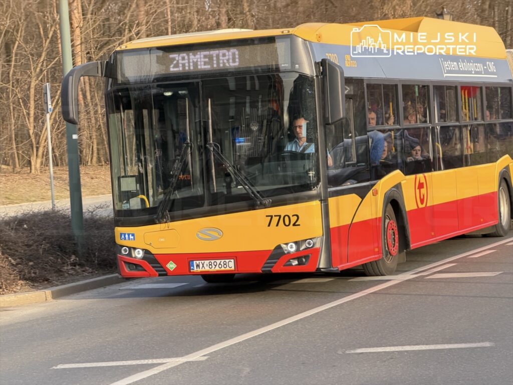 Autobus miejski z numerem 7702 z napisem "ZAMETRO" przejeżdża obok przystanku w otoczeniu drzew.