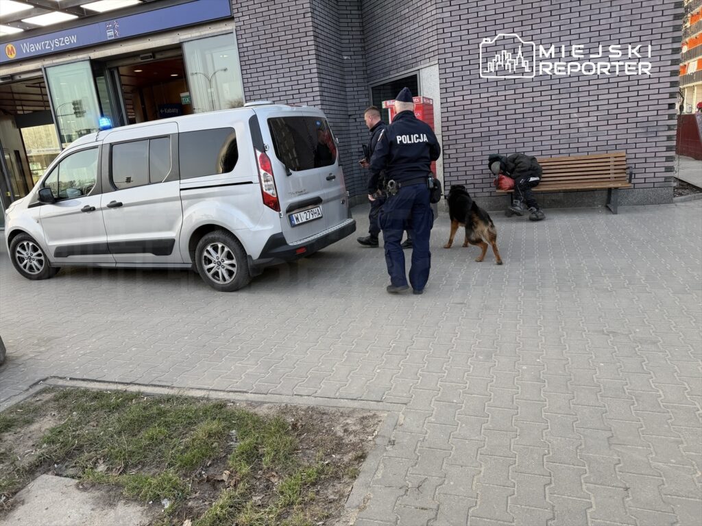 Policjanci z psem służbowym przeprowadzają interwencję przy przystanku metra, obok zaparkowanego samochodu.