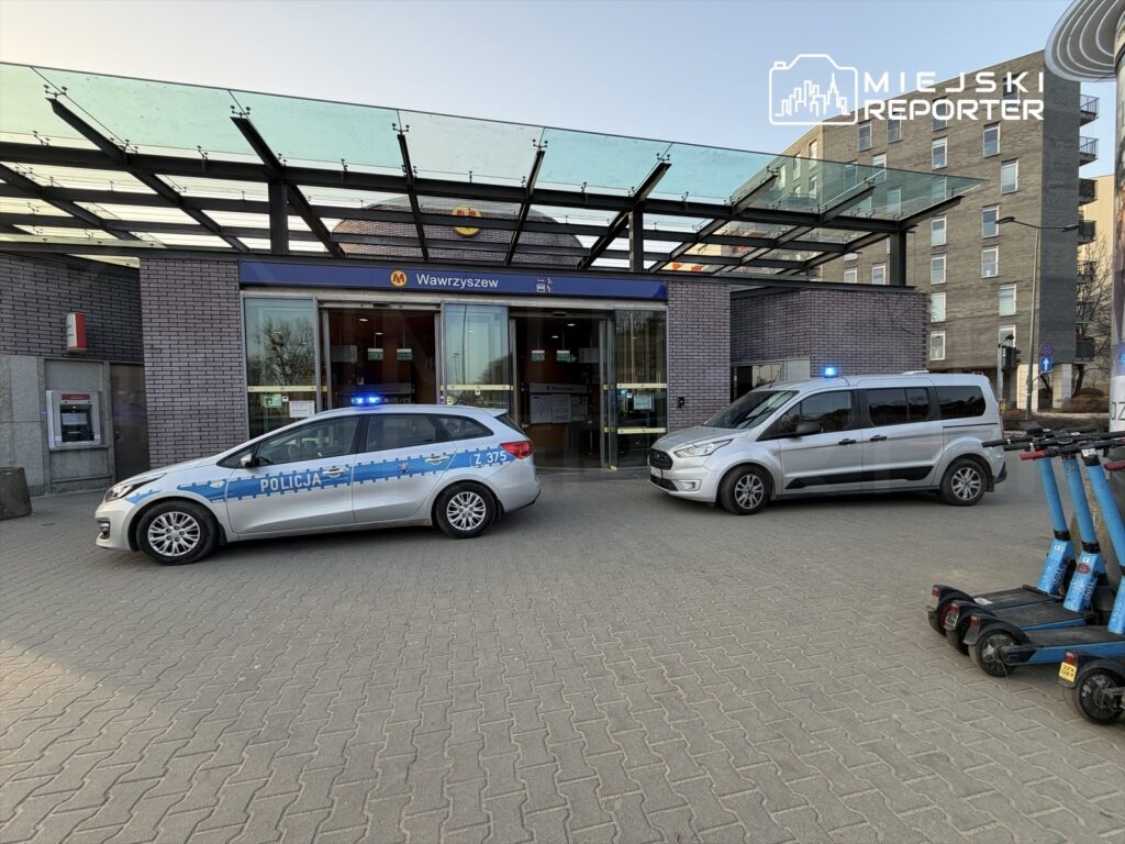 Policyjny radiowóz z niebieskimi światłami zaparkowany przed wejściem do stacji metra, obok drugiego pojazdu policyjnego.