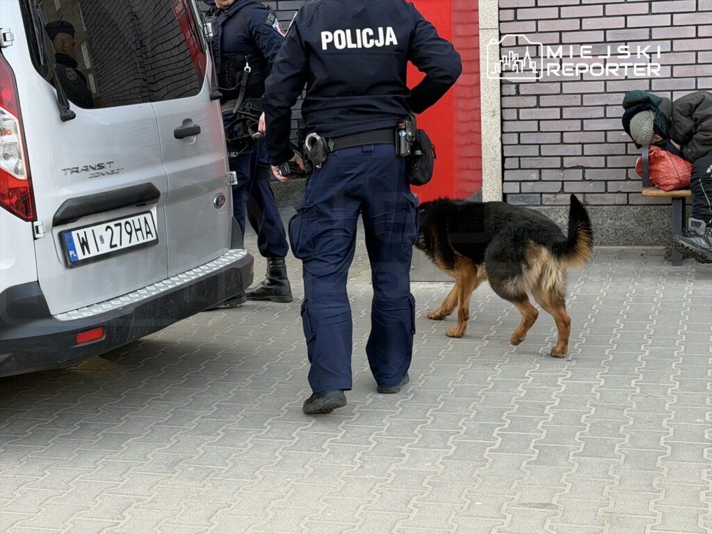 Policjant z psem tropiącym bada teren obok białego furgonu zaparkowanego na chodniku.