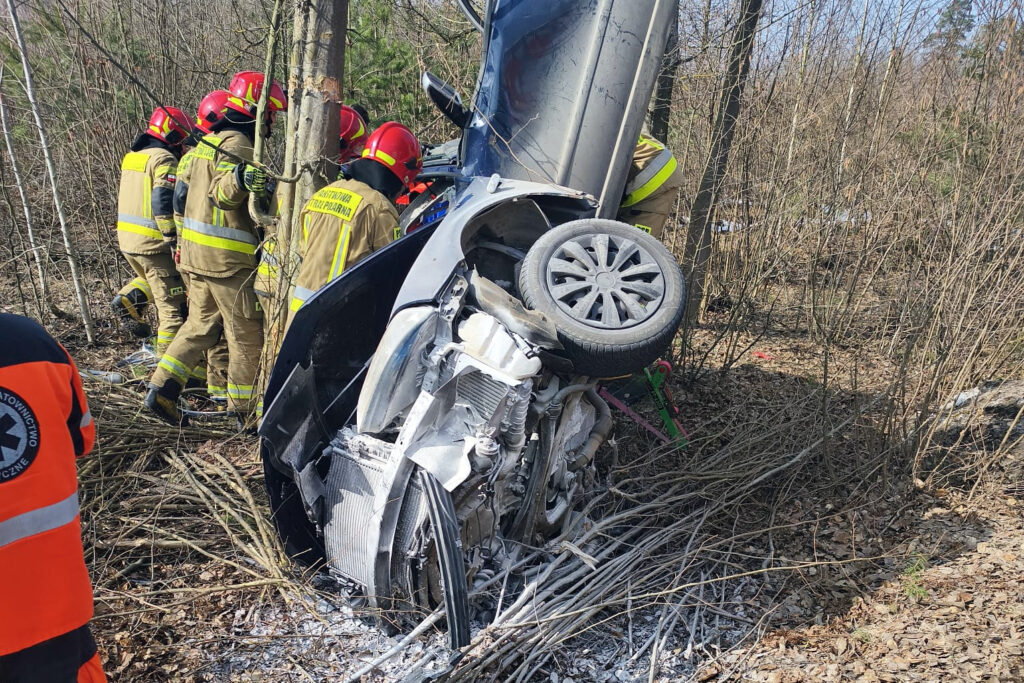 Grupa strażaków w hełmach pracuje nad przewróconym samochodem w lesie, otoczonym gałęziami i krzakami.