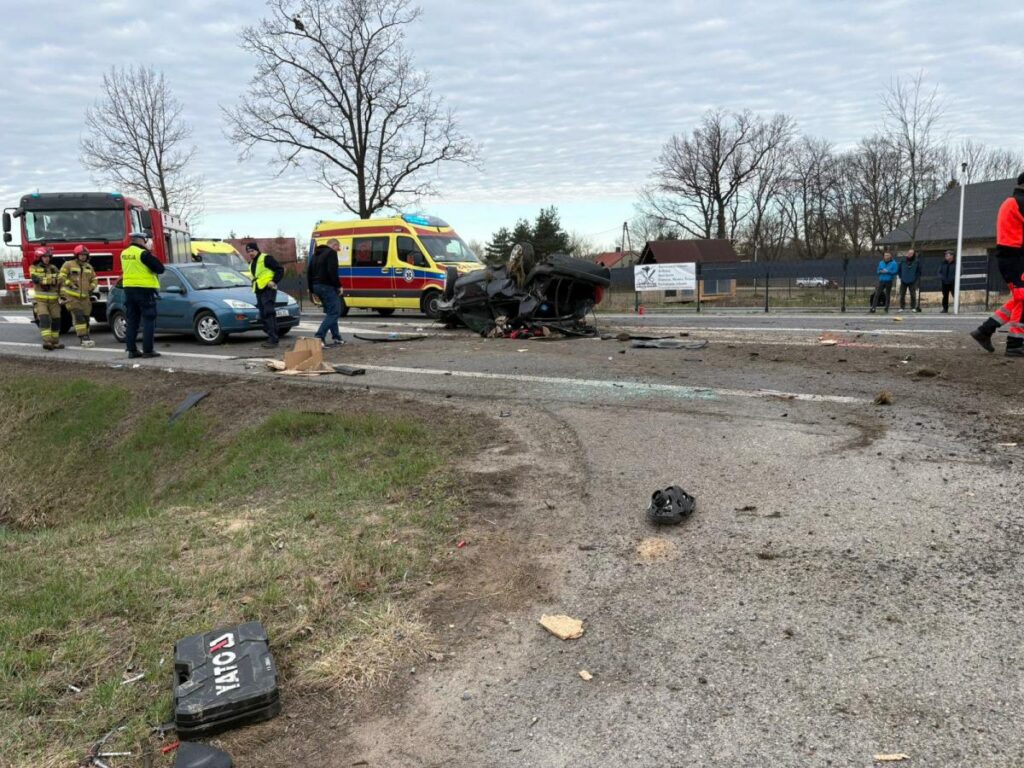 Na drodze leżą wrak samochodu oraz rozbite elementy, obok pracują strażacy i ratownicy medyczni.