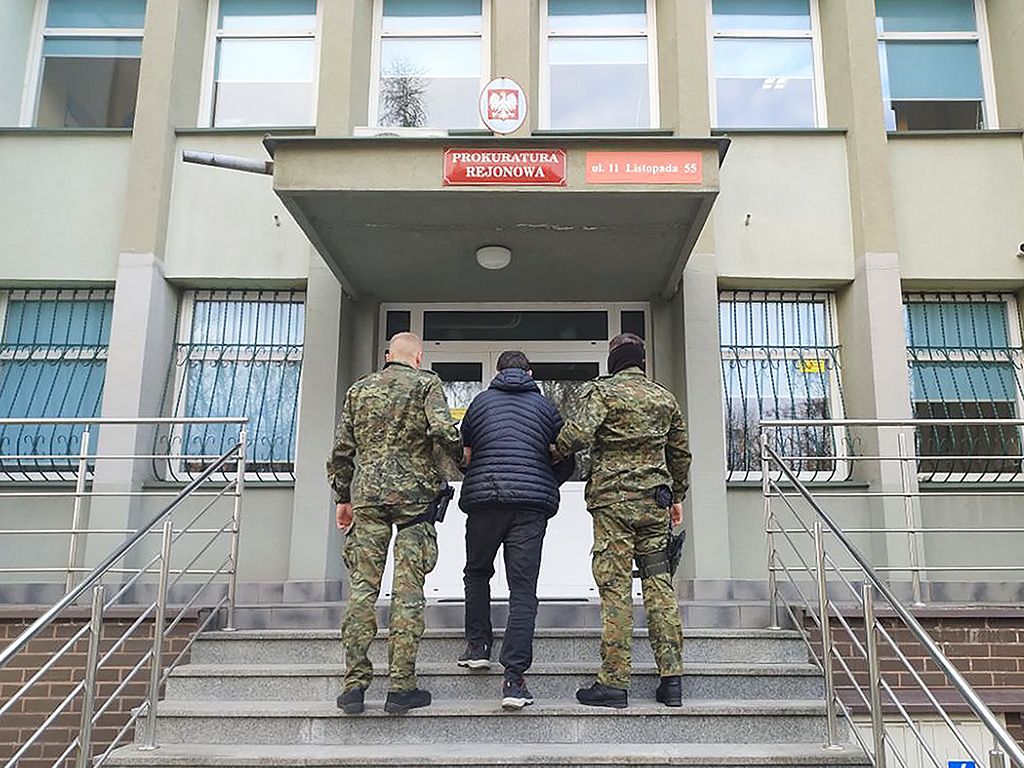 Mężczyzna w czarnej kurtce prowadzony przez dwóch funkcjonariuszy w mundurach do prokuratury.